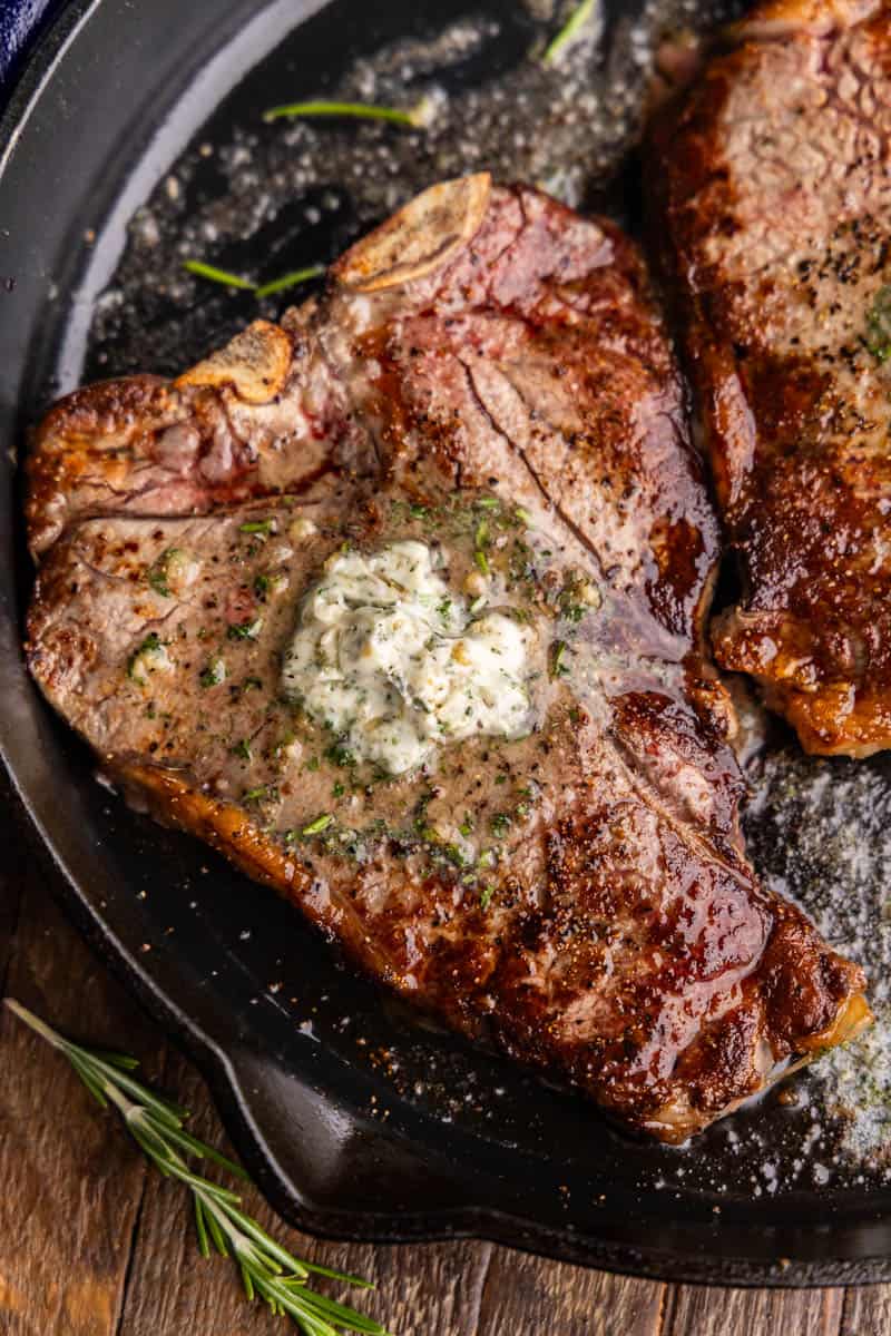 An overhead view of a t-bone steak in a cast iron skillet with resting butter melting on top.