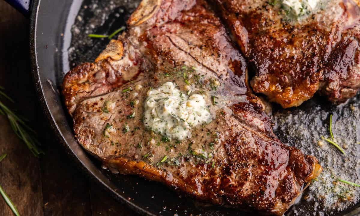 A closeup overhead view of a t-bone steak in a cast iron skillet with resting butter melting on top.
