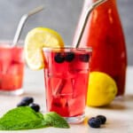 Glass of Blueberry Lemonade with a pitcher in the background.