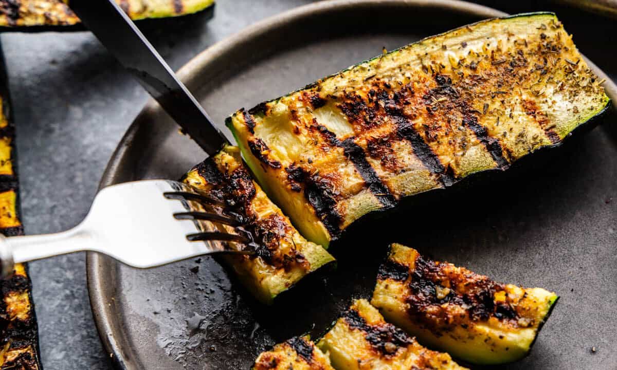 A grilled piece of zucchini is cut with a knife and fork.