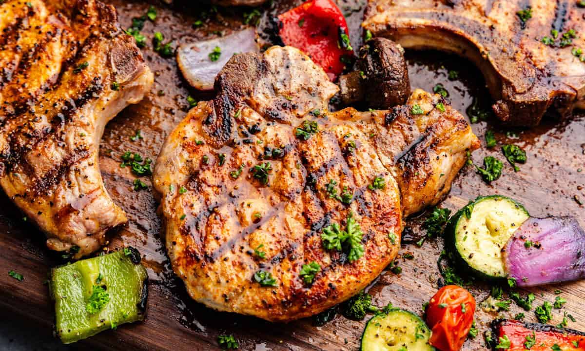 Grilled pork chops on a wooden board with grilled vegetables around them.