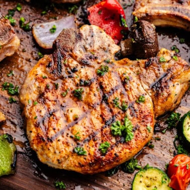 Grilled pork chops on a wooden board with grilled vegetables around them.