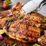 A grilled pork chop being lifted from a wood board with a pair of tongs.