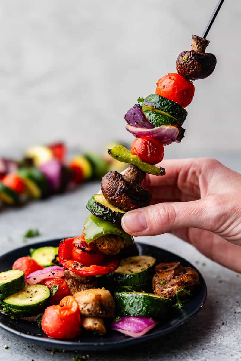 One kabob being held up above a plate of grilled vegetables.