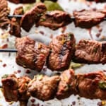 Three grilled beef kebabs with spices on the table underneath.