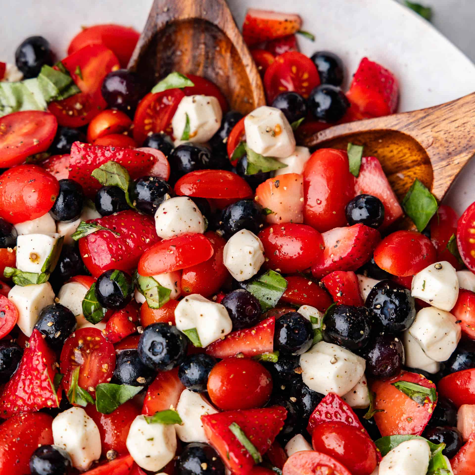 A close view of a bowl of red, white, and blue caprese salad with a wooden spoon.