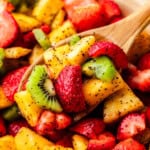 A close up view of a wooden spoon dishing out a helping of summer fruit salad with pineapple, kiwi, and strawberry.