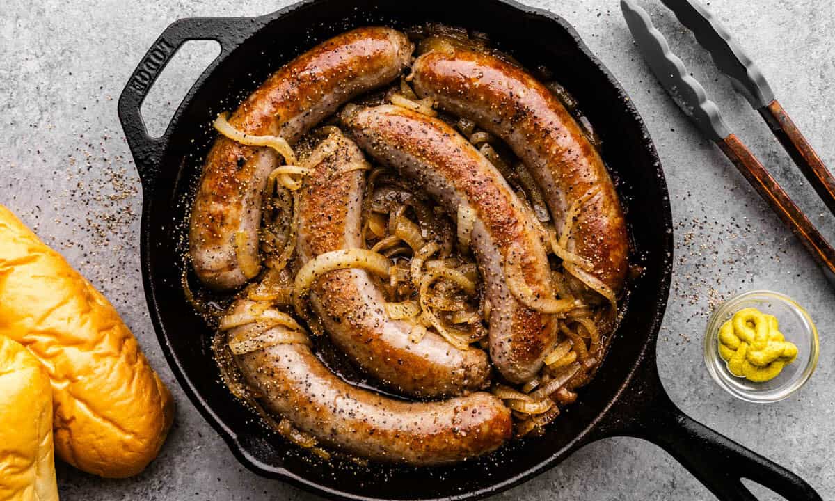 An overhead view of a cast iron skillet filled with 5 bratwurst sausages braised in beer and onions.