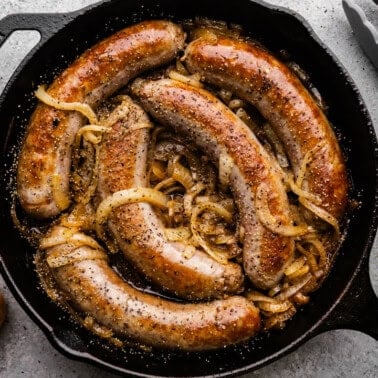 An overhead view of a cast iron skillet filled with 5 bratwurst sausages braised in beer and onions.