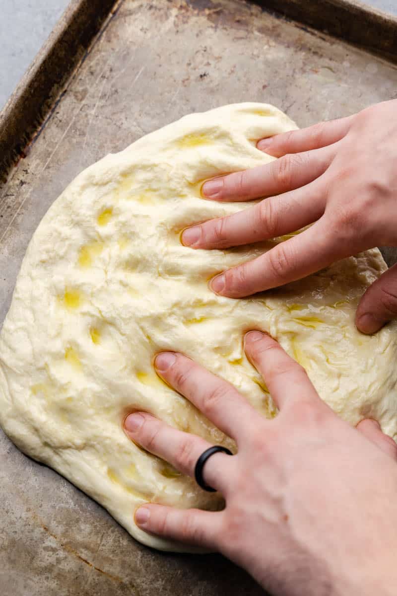 Hands shaping a loaf of focaccia, making indents in the dough.