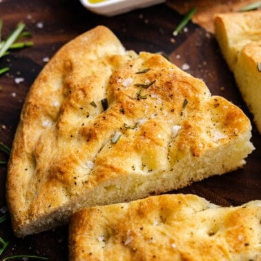 A loaf of homemade rosemary focaccia that has been cut into wedges. It is sitting on a wooden cutting board.