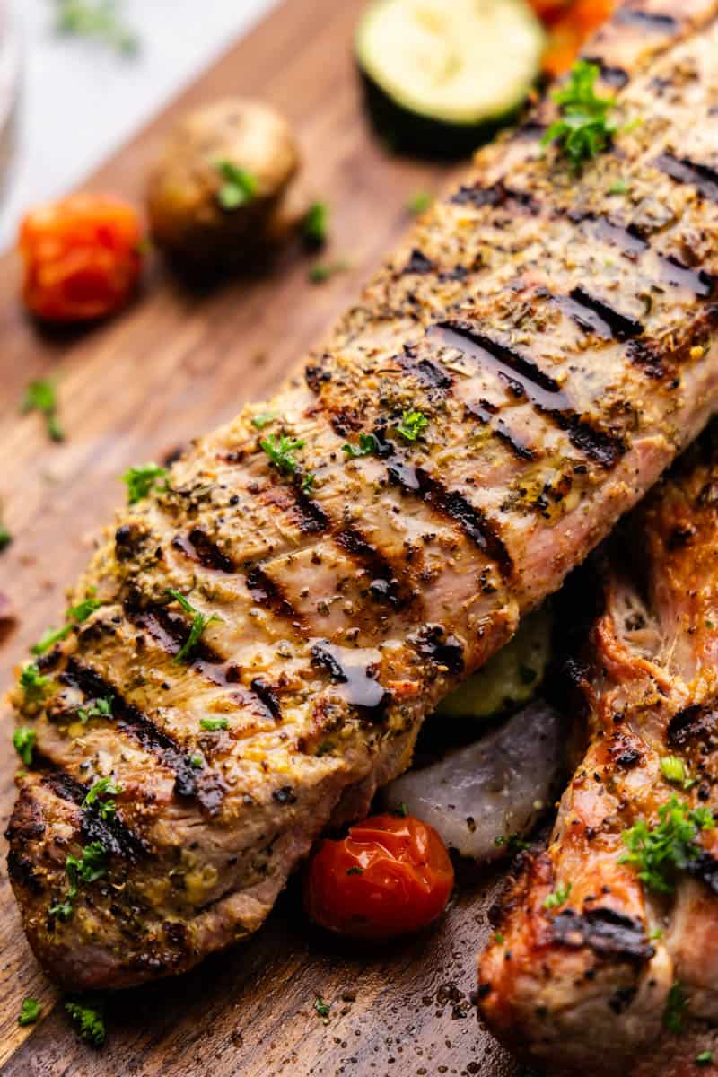 An entire grilled pork tenderloin is laid out on a wooden cutting board.
