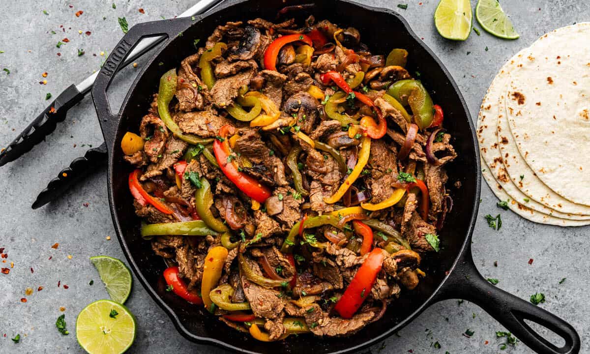 An overhead view of a cast iron skillet filled with steak fajitas, sitting on a counter next to flour tortillas and tongs.