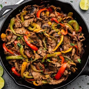 An overhead view of a cast iron skillet filled with steak fajitas, sitting on a counter next to flour tortillas and tongs.