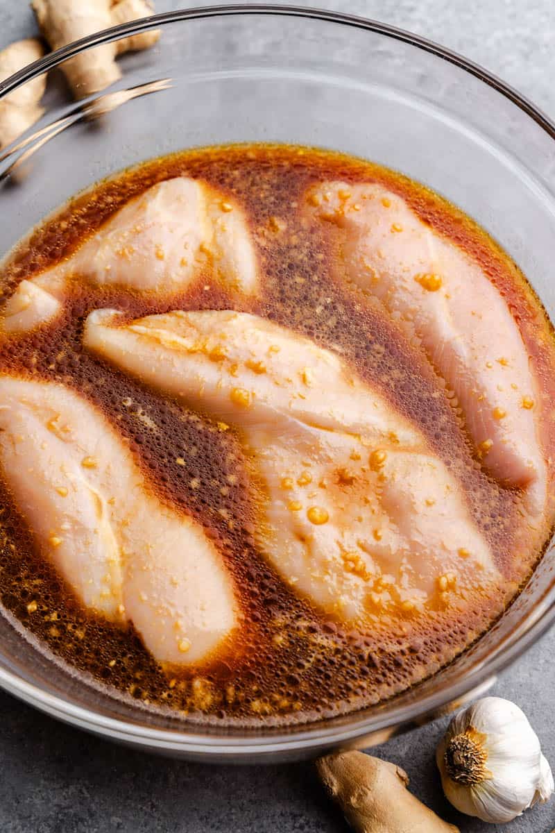 Raw chicken breasts soaking in a bowl of reddish brown marinade.