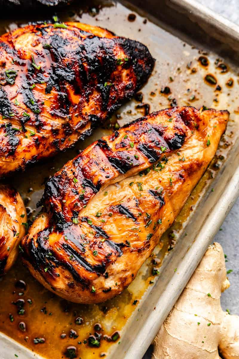 A closeup overhead photo of a grilled golden chicken breast on a baking sheet with seasonings and other chicken breasts on the side.