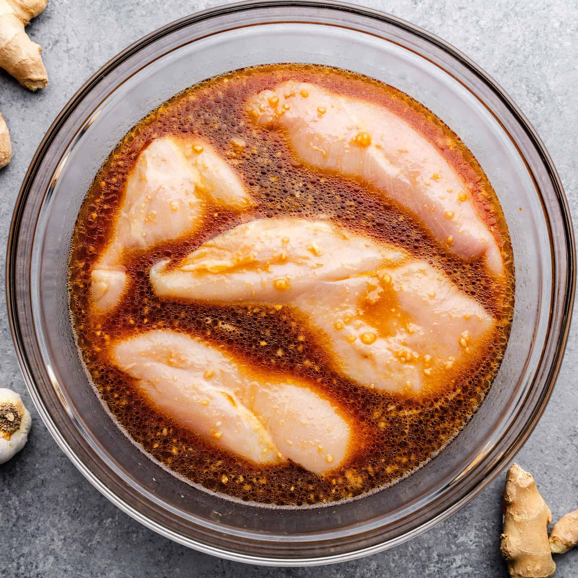 An overhead picture of a glass mixing bowl filled with raw chicken breasts and a reddish brown marinade sauce.