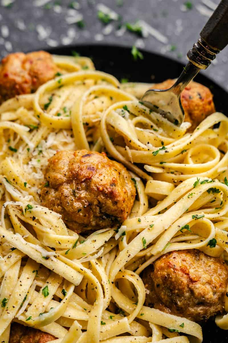 Fettuccini alfredo with baked chicken meatballs placed on top, with a fork digging in for a bite.