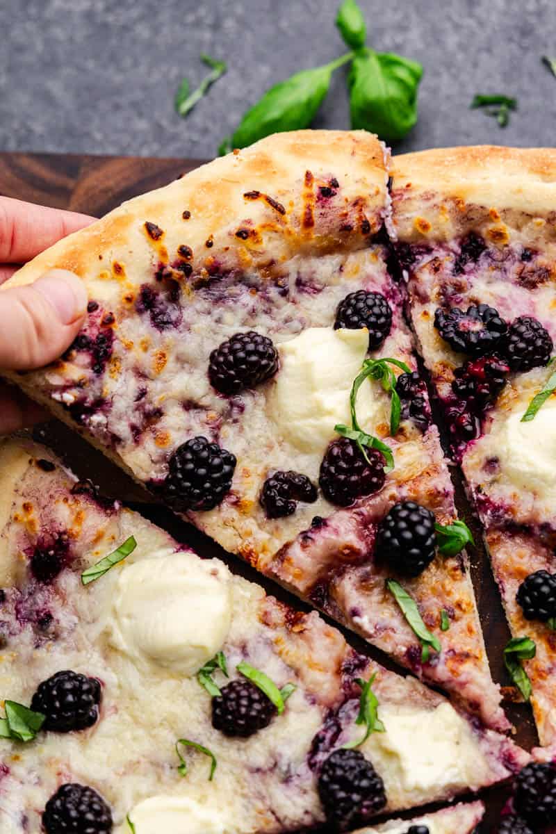 A close up of a slice of a blackberry basil ricotta pizza slice being taken from the rest of the pie.