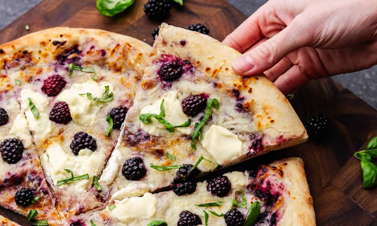 An overhead shot of a slice of blackberry basil ricotta pizza being lifted by a hand away from the rest of the pizza.