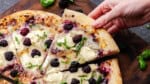 An overhead shot of a slice of blackberry basil ricotta pizza being lifted by a hand away from the rest of the pizza.