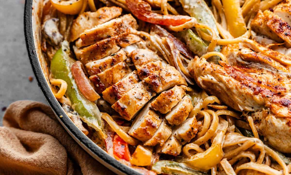 An overhead view of a skillet filled with browned sliced chicken breast, noodles and bell peppers.