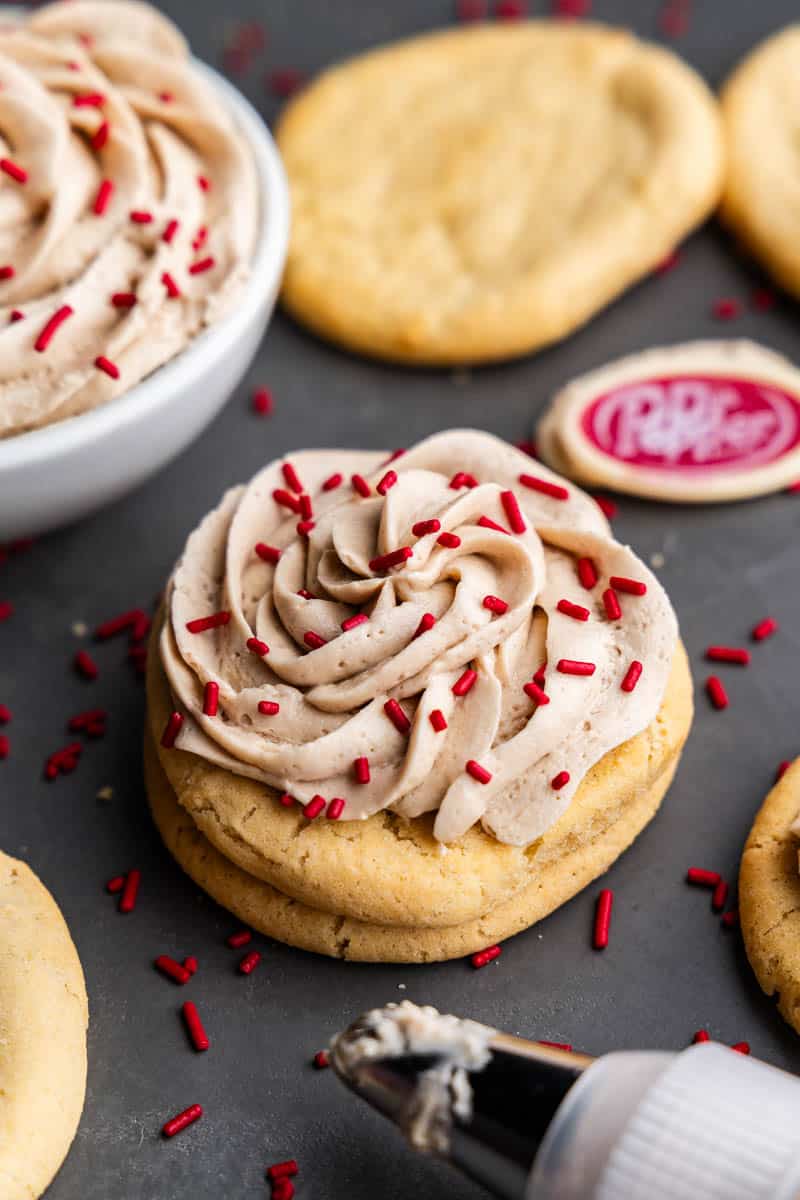 An angled view of a sugar cookie topped with a swirl of Dr. Pepper frosting and red sprinkles, with a small Dr. Pepper logo in the background.