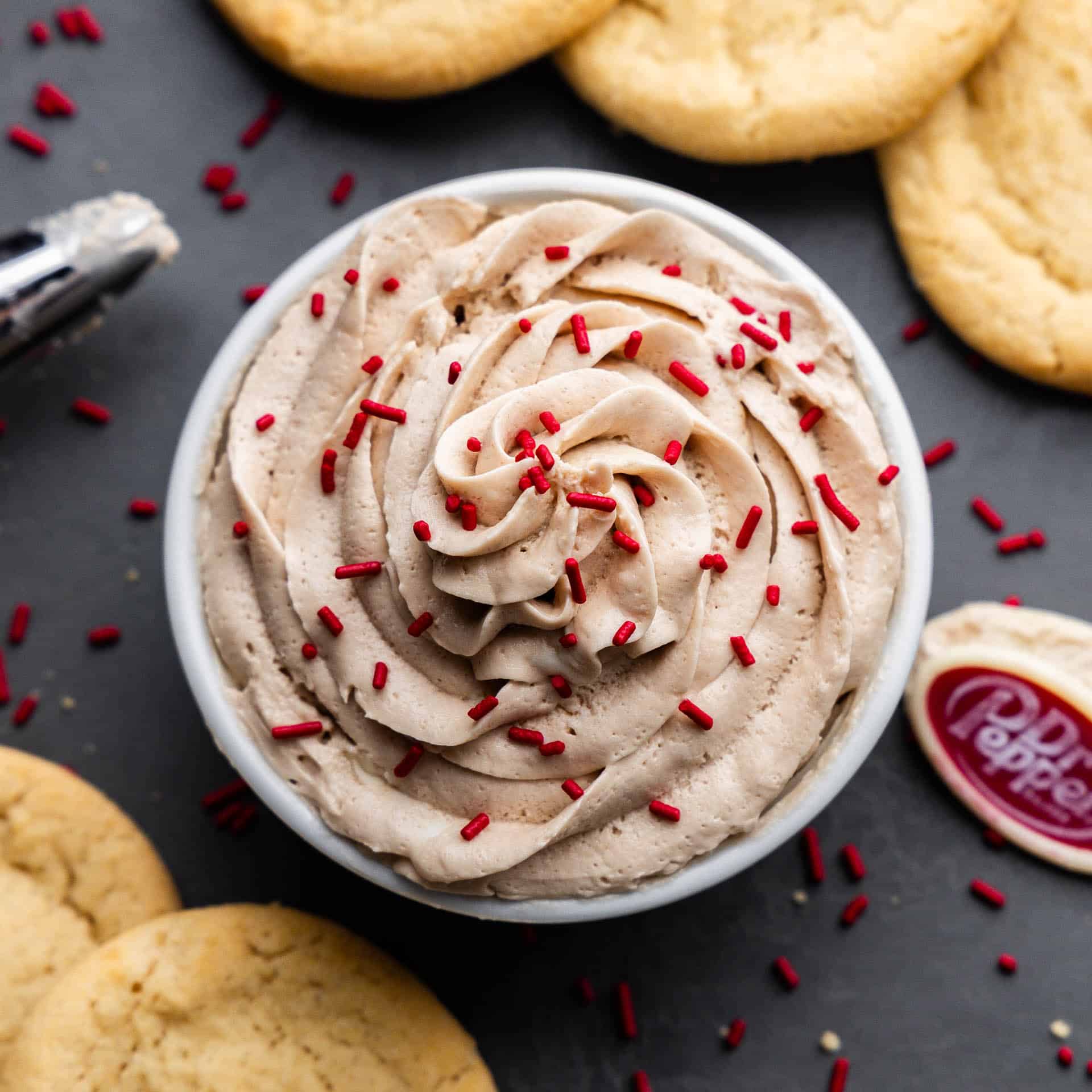 An overhead view of a bowl of Dr. Pepper frosting topped with red sprinkles and surrounded by unfrosted sugar cookies.