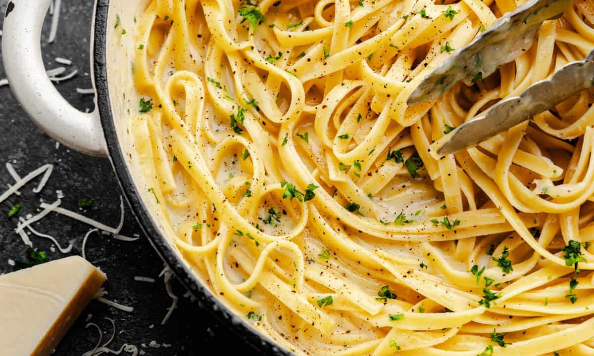 A close up overhead shot of a pot filled with fettuccine alfredo, with tongs set inside to serve.