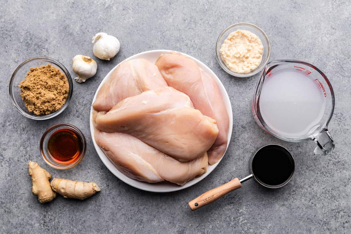 An overhead image of a plate of raw chicken breast surrounded by cups and spoons filled with wet and dry ingredients.