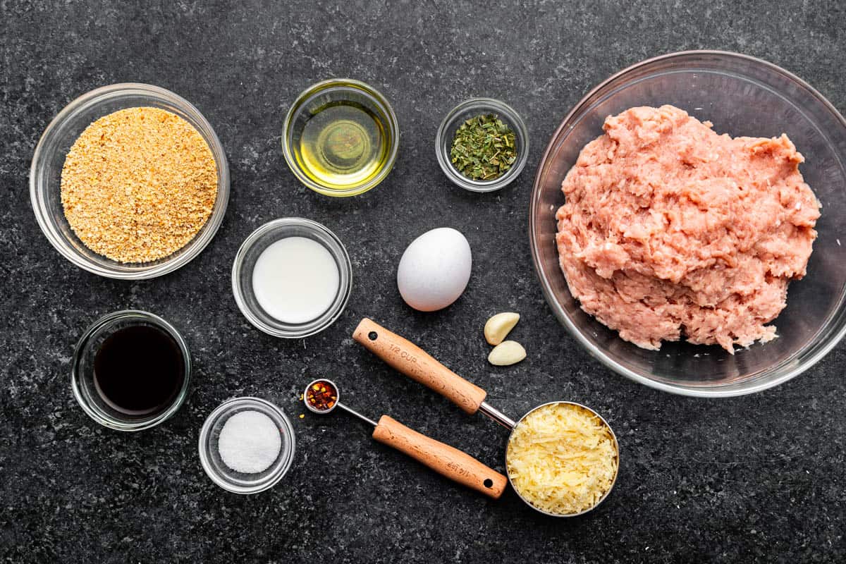 A bowl of raw ground chicken, surrounded by other bowls of measured ingredients, an egg and measuring spoons with ingredients.