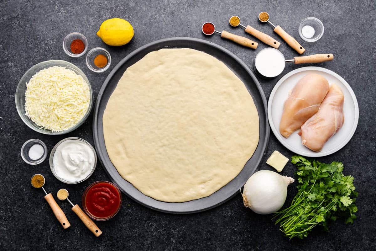 An overhead shot of raw pizza dough rolled out, next to a plate of raw chicken breasts, surrounded by measuring spoons and cups filled with ingredients and raw vegetables.