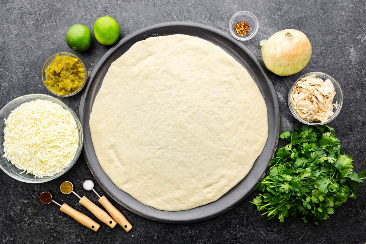 An overhead photo of raw pizza dough rolled out on a circular baking sheet, surrounded by fresh ingredients and measured dry and wet ingredients.