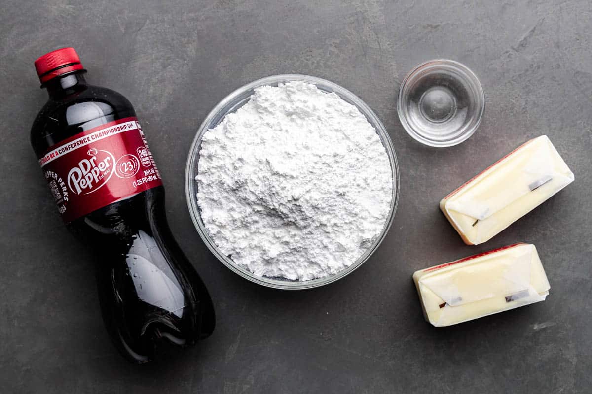 An overhead view of a bowl of powdered sugar, a soda bottle of Dr. Pepper, sticks of butter and a small bowl of vanilla extract.