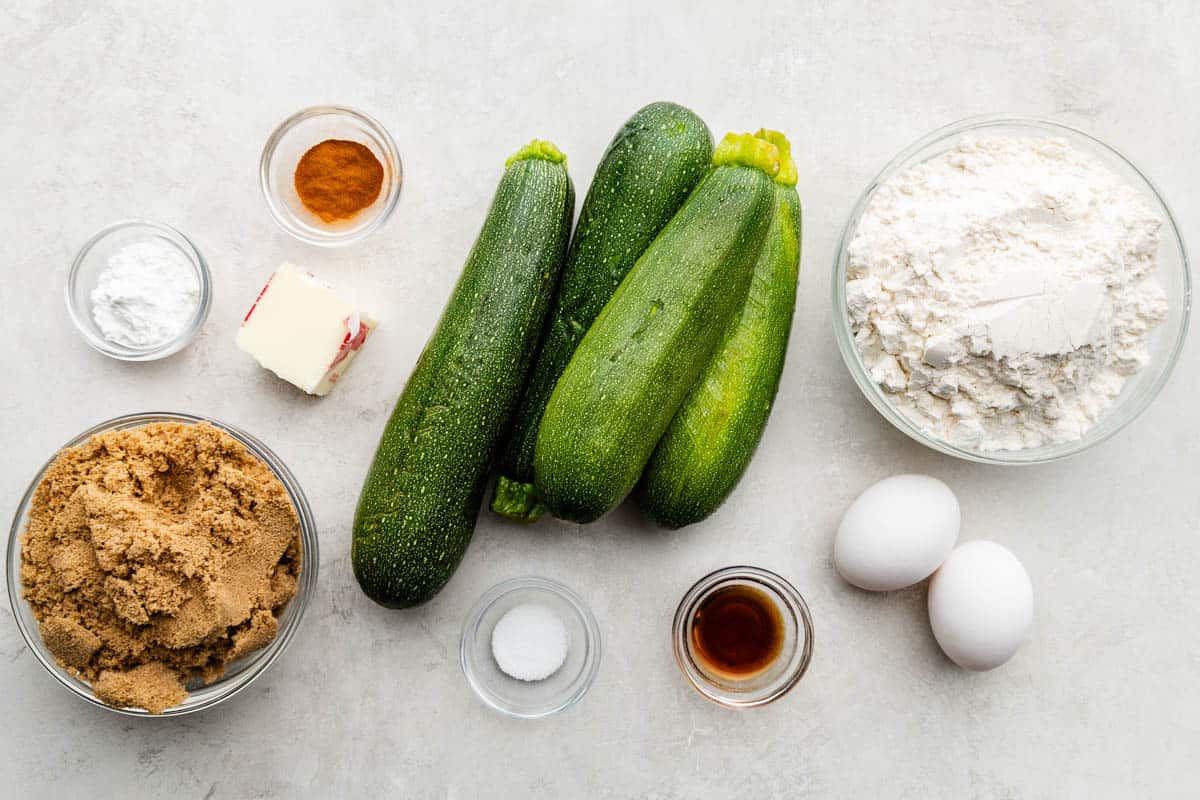 An overhead shot of several whole zucchinis surrounded by wet and dry ingredients and a bowl of whole eggs.