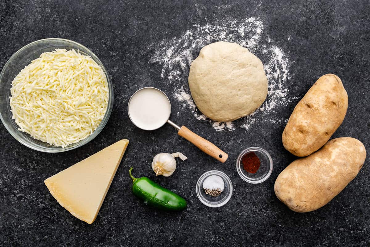 A ball of raw pizza dough sitting between whole potatoes, a cheese wedge, a jalapeño, a bowl of shredded cheese and a measuring cup with other ingredients.