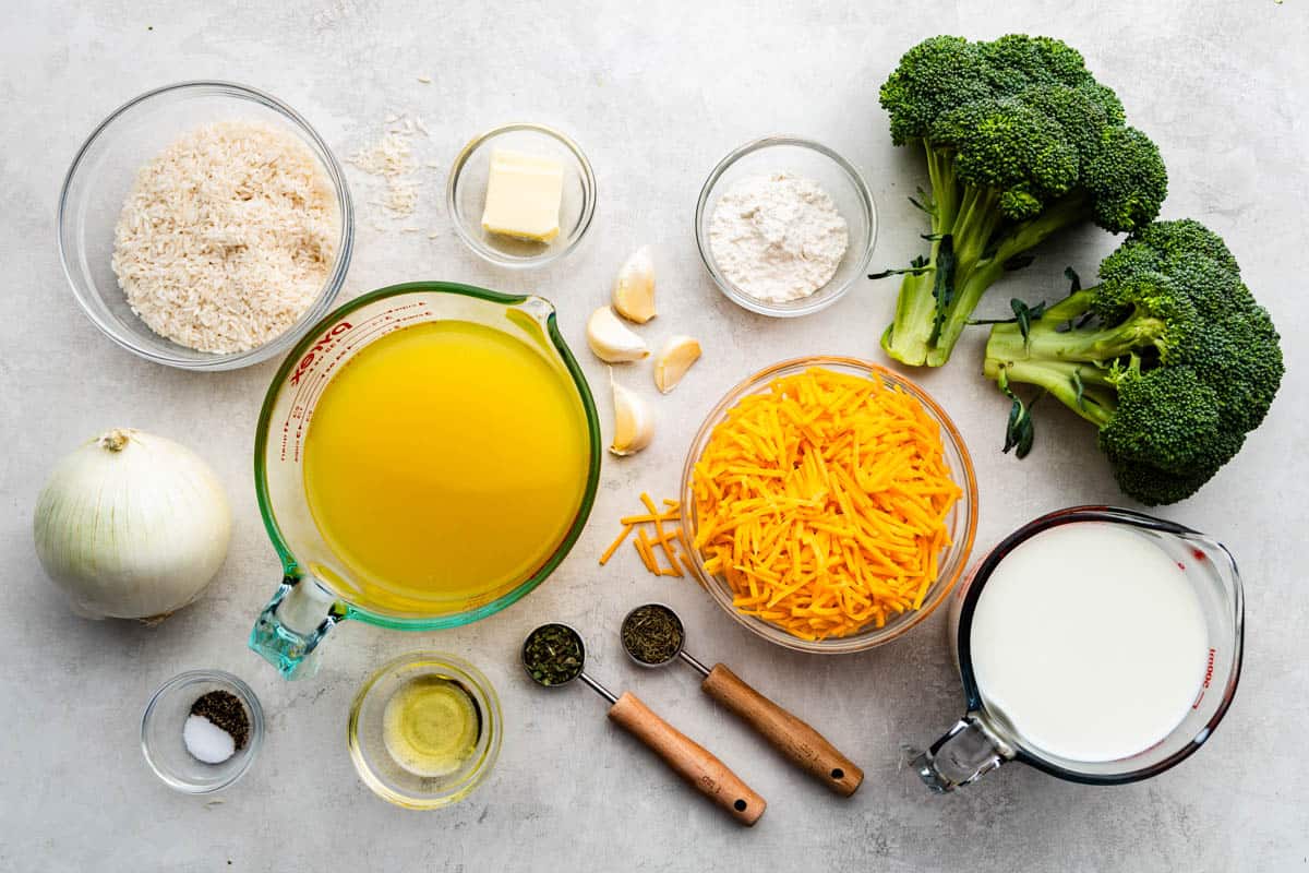 An overhead shot of raw and dried ingredients, including heads of broccoli, a bowl of shredded cheddar cheese, and measuring cups and spoons with ingredients.