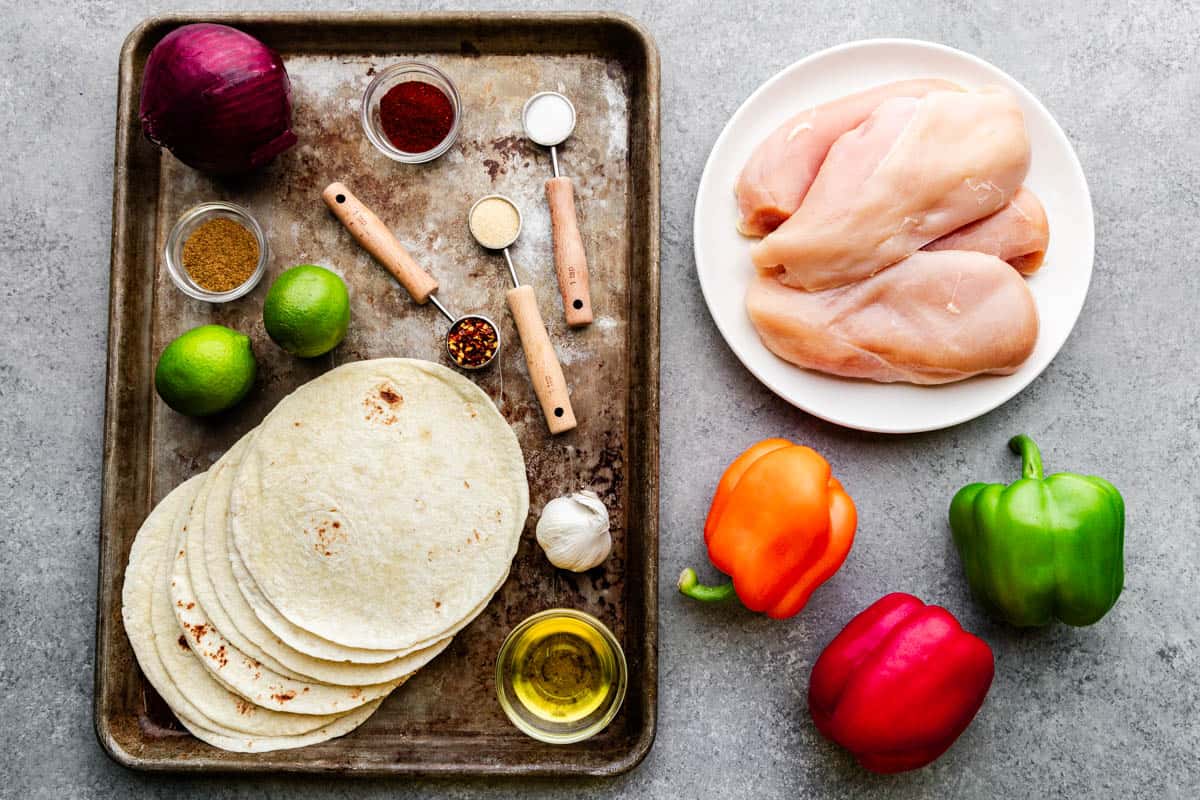 A sheet pan filled with flour tortillas, whole limes and measured spices, a plate of raw chicken and three whole bell peppers.