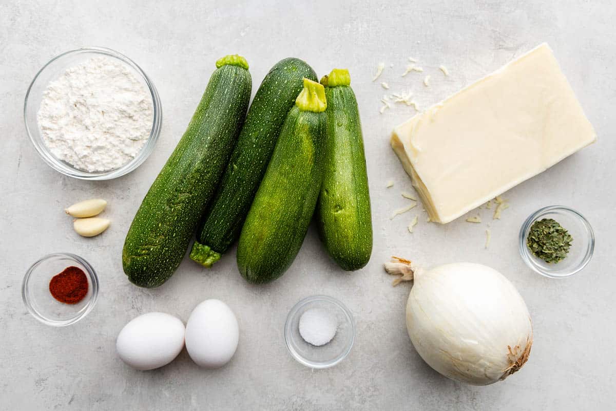 An overhead picture of whole zucchinis, fresh vegetables and a chunk of cheese with eggs and dry and wet ingredients.