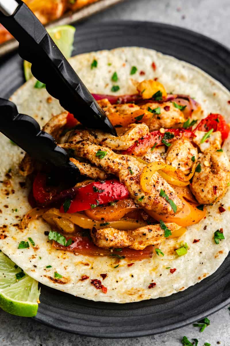 Tongs placing chicken fajitas onto a flour tortilla.