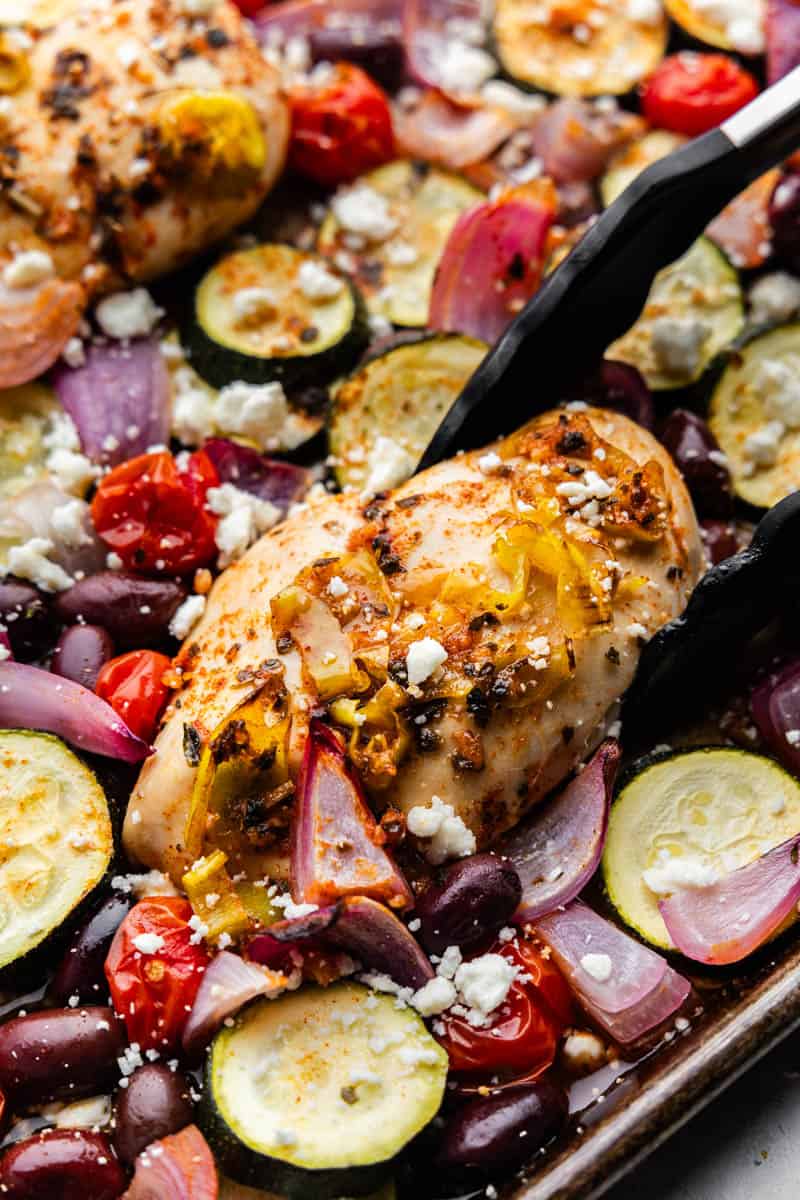 Tongs lifting a piece of greek chicken off a sheet pan of roasted vegetables.