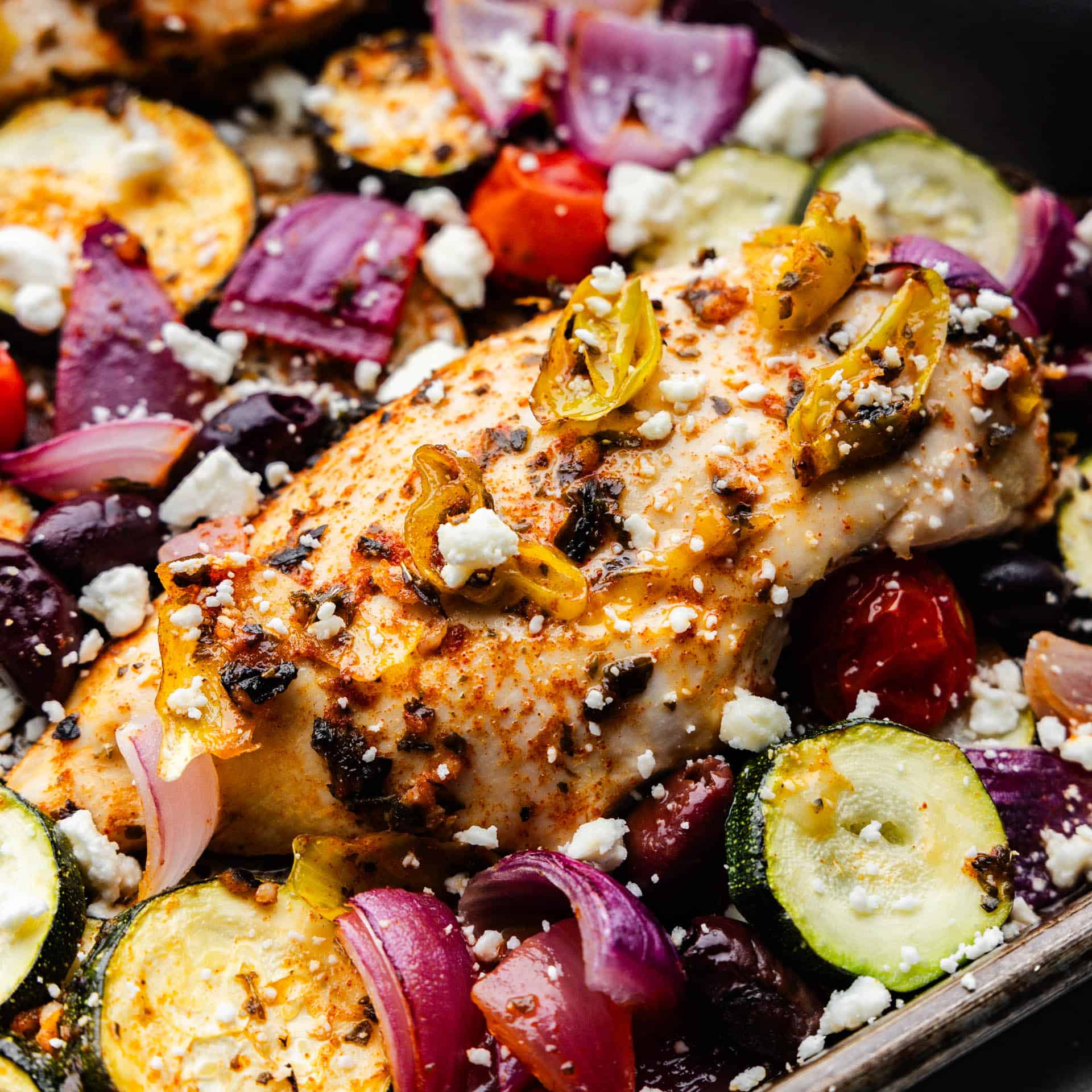 A close up view of greek chicken surrounded by vegetables on a sheet pan.