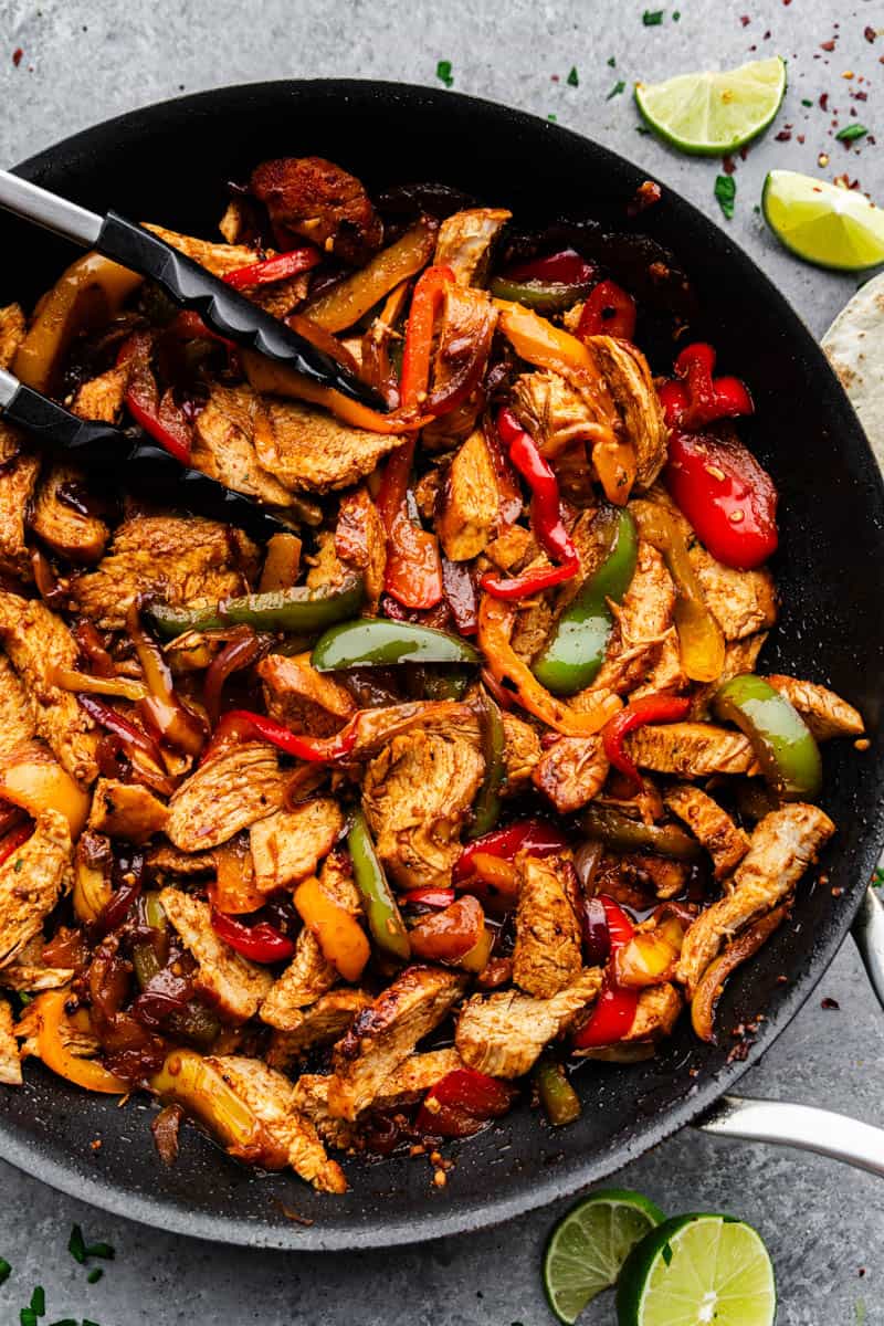 An overhead close up view of a skillet filled with chicken fajitas, with tongs placed in the skillet.