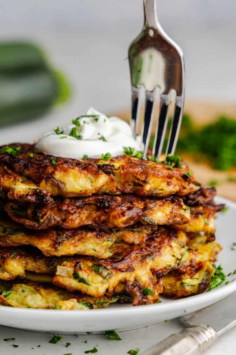 A side picture of a stack of zucchini pancakes topped with a dollop of sour cream, with a fork hovering over the top to take a bite.