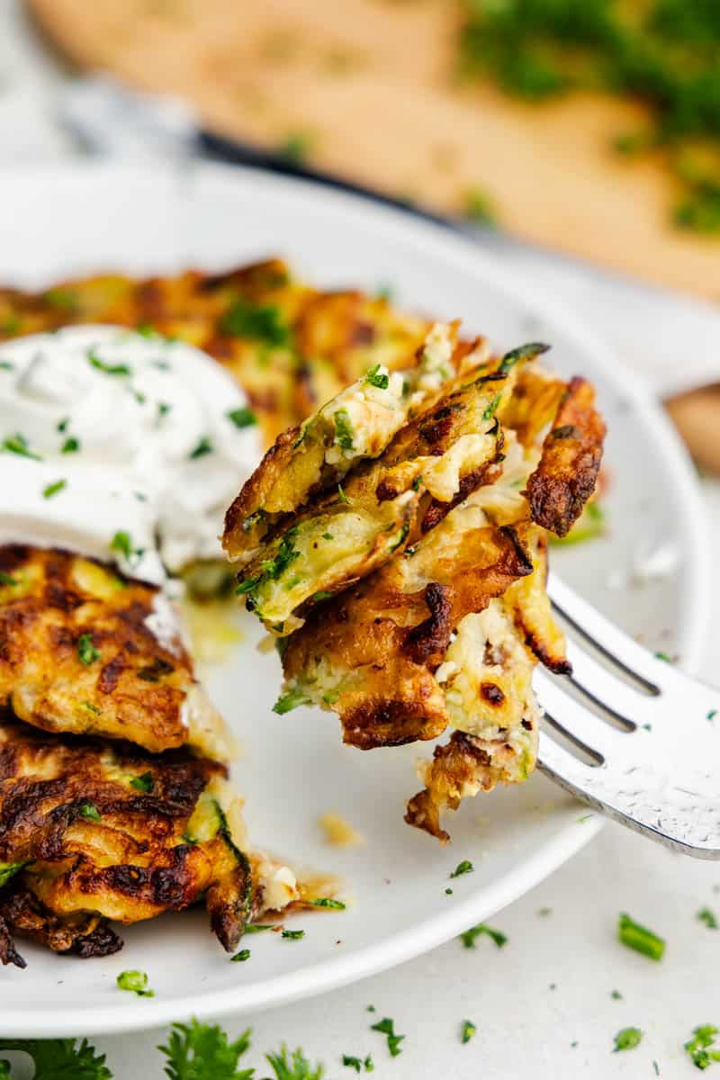 A bite of zucchini pancakes on a fork against a plate with the stack of zucchini pancakes.