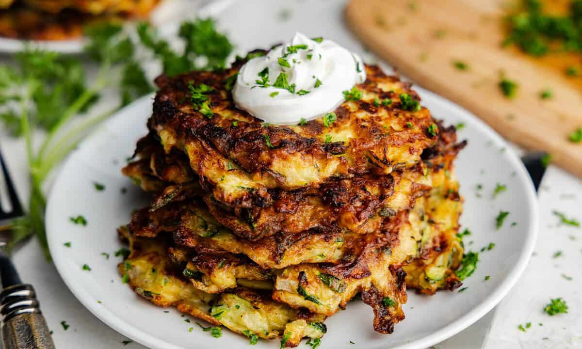 A stack of crispy brown zucchini pancakes on a plate topped with a dollop of sour cream and fresh herbs.