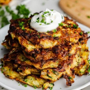 A stack of crispy brown zucchini pancakes on a plate topped with a dollop of sour cream and fresh herbs.