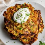 A top image of a stack of crispy brown zucchini pancakes on a white plate topped with sour cream and fresh green herbs.