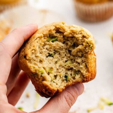 A hand holding a zucchini muffin with a large bite taken out of it.