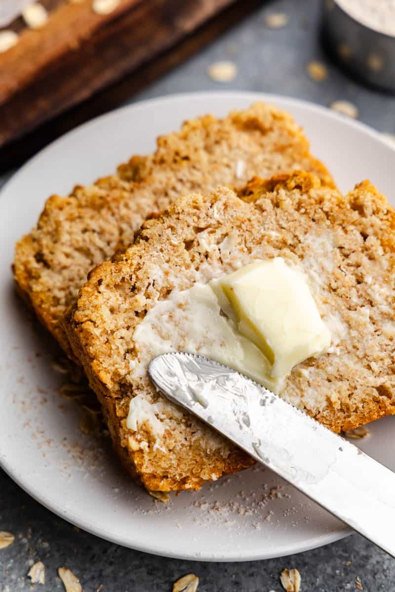 Two slices of beer bread on a plate with a knife spreading butter on top.